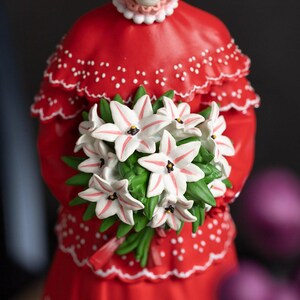 Puede incluir: Figura roja de una mujer sosteniendo un ramo de lirios blancos con rayas rosas. El vestido de la mujer es rojo con lunares blancos y un ribete con volantes. Las flores tienen hojas verdes.