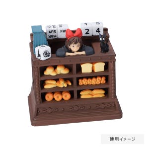 Pode incluir: Uma exibi&ccedil;&atilde;o de padaria em miniatura marrom com uma jovem com um la&ccedil;o vermelho, um gato preto e v&aacute;rios produtos de panifica&ccedil;&atilde;o. A exibi&ccedil;&atilde;o inclui um calend&aacute;rio com a data 24 e o texto "APR FRI".