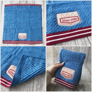 Puede incluir: Toalla de mano azul con un patr&oacute;n texturizado y un borde a rayas rojo y blanco. La toalla presenta un parche rectangular con el texto "Since 1988" y un gr&aacute;fico de tres p&aacute;jaros.