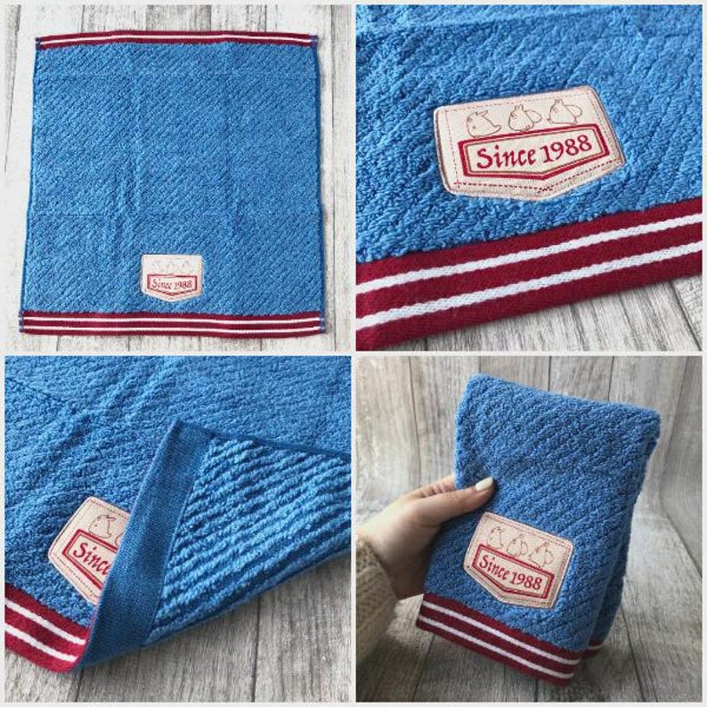 Puede incluir: Toalla de mano azul con un patr&oacute;n texturizado y un borde a rayas rojo y blanco. La toalla presenta un parche rectangular con el texto "Since 1988" y un gr&aacute;fico de tres p&aacute;jaros.