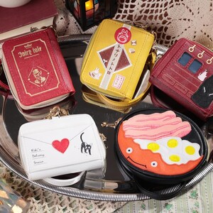 May include: A collection of small, zippered wallets with unique designs, including a red book-shaped wallet, a yellow wallet with Japanese text, and a red wallet with a character illustration. The wallets are arranged on a silver tray.