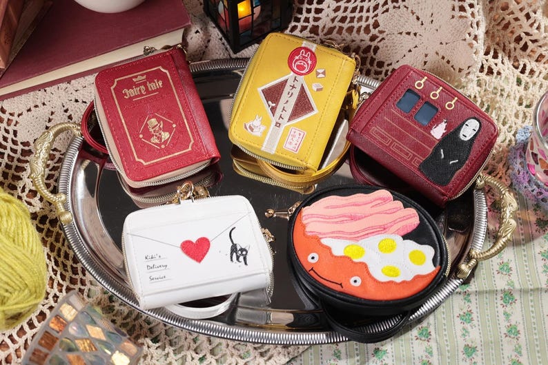 May include: A collection of small, zippered wallets with unique designs, including a red book-shaped wallet, a yellow wallet with Japanese text, and a red wallet with a character illustration. The wallets are arranged on a silver tray.