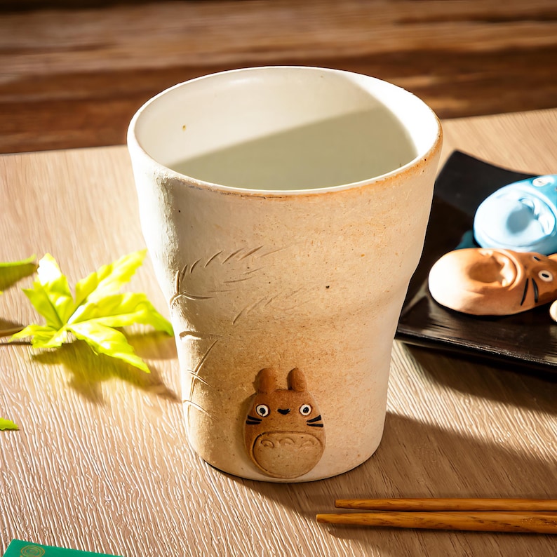 May include: A beige ceramic mug with a Totoro character design. The mug has a cylindrical shape with a slightly flared top and a textured surface. The Totoro figure is brown with black details. The mug is on a wooden surface.