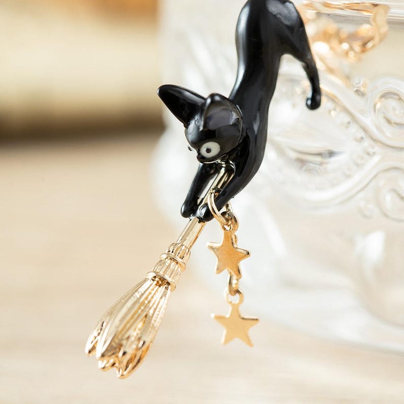 May include: A black cat-shaped brooch with a gold-colored broom and dangling star charms. The cat is reaching down towards the broom, with its eyes wide open. The brooch has a glossy finish and is set against a blurred background.