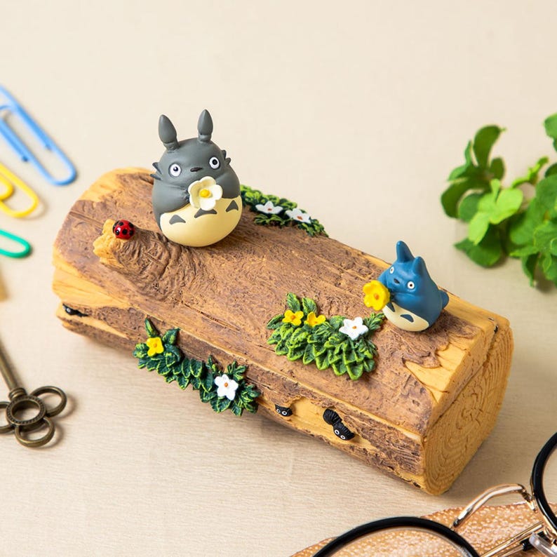 May include: A decorative log-shaped box featuring two Totoro figurines, one gray and one blue, each holding a flower. The box is adorned with miniature flowers, greenery, and a ladybug. The box is made of a brown material.