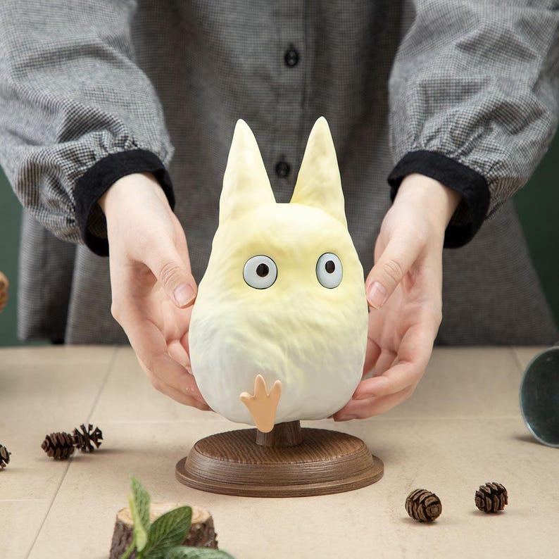 May include: A light yellow, cartoon-style character with large blue eyes and pointed ears, mounted on a brown wooden base. The character has a small, orange-colored foot. The figure is held by a person, with a neutral background.