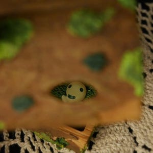 Pode incluir: Uma pequena figura do esp&iacute;rito Kodama, esbranqui&ccedil;ada, espreitando de uma base castanha texturizada com detalhes verdes. A figura tem um rosto sorridente e &eacute; rodeada por folhagem verde. A base tem um aspeto de madeira.