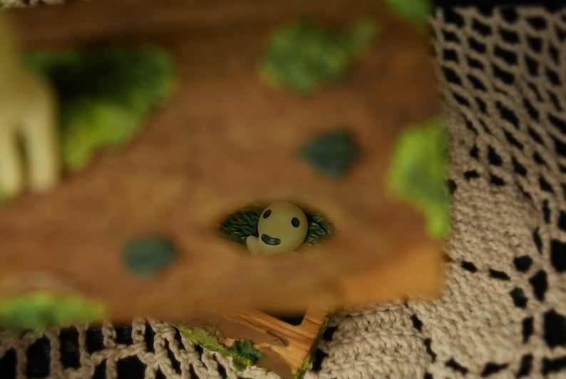 Pode incluir: Uma pequena figura do esp&iacute;rito Kodama, esbranqui&ccedil;ada, espreitando de uma base castanha texturizada com detalhes verdes. A figura tem um rosto sorridente e &eacute; rodeada por folhagem verde. A base tem um aspeto de madeira.