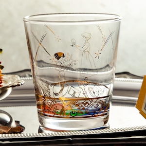 May include: A clear glass tumbler with a gold and black illustration of two figures and a small creature. The design includes gold accents and a colorful band at the base. The glass is set on a silver tray.