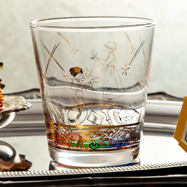 May include: A clear glass tumbler with a gold and black illustration of two figures and a small creature. The design includes gold accents and a colorful band at the base. The glass is set on a silver tray.