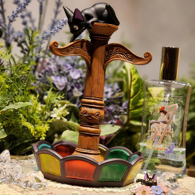 May include: A decorative, wooden cat-themed perfume stand with a stained glass base. The stand features a black cat figurine perched atop a brown, ornate post. A perfume bottle is visible in the background.