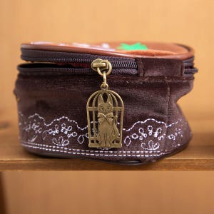 May include: A brown velvet cosmetic bag with a decorative cat charm. The bag features a zipper closure and a white embroidered floral design along the bottom. The top of the bag has a brown and green design.