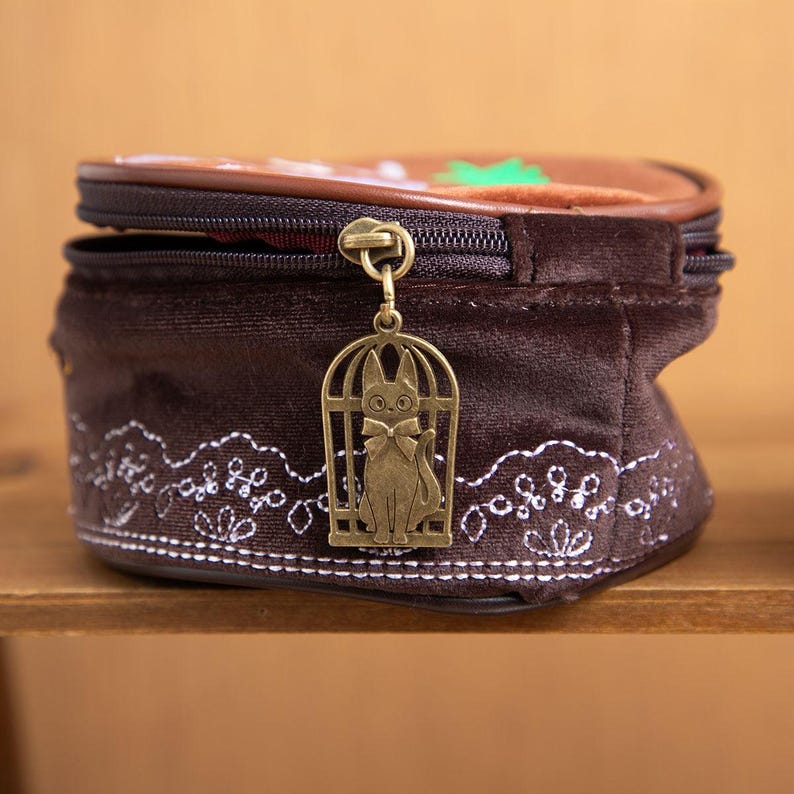 May include: A brown velvet cosmetic bag with a decorative cat charm. The bag features a zipper closure and a white embroidered floral design along the bottom. The top of the bag has a brown and green design.