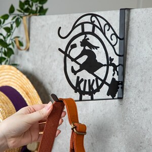 K&ouml;nnte beinhalten: Schwarzer Metall-T&uuml;rhaken mit einer Silhouette einer Hexe auf einem Besen, darunter das Wort "KIKI". Der Haken h&auml;lt einen braunen und orangefarbenen Taschenriemen. Im Hintergrund ist ein Strohhut zu sehen.