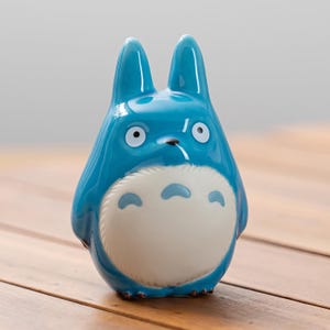 May include: A glossy, blue ceramic figurine of a Totoro character. The figure has large ears, white eyes, and a white belly with blue markings. The figurine is set against a wooden surface.