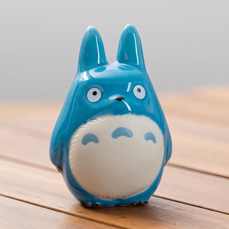 May include: A glossy, blue ceramic figurine of a Totoro character. The figure has large ears, white eyes, and a white belly with blue markings. The figurine is set against a wooden surface.