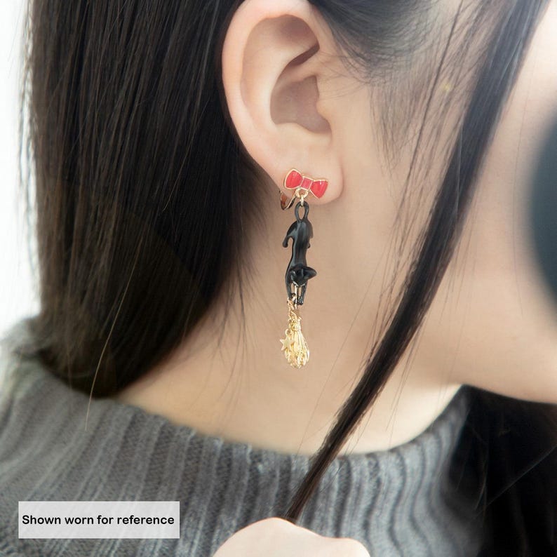 May include: Black cat earrings with a red bow accent. The earrings feature a black cat hanging from a gold-colored broom. The earrings are shown worn for reference.