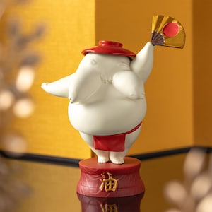 Peut inclure: Figurine blanche repr&eacute;sentant un lutteur de sumo avec un chapeau et un pagne rouges, tenant un &eacute;ventail dor&eacute; avec un cercle rouge. La figurine est sur une base rouge avec des caract&egrave;res japonais.