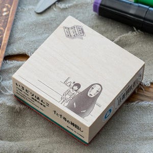 May include: A square wooden stamp featuring a black ink illustration of the characters Chihiro and No-Face from Spirited Away. The stamp includes Japanese text, the code TSW-194, and Studio Ghibli branding printed on the light wood surface.