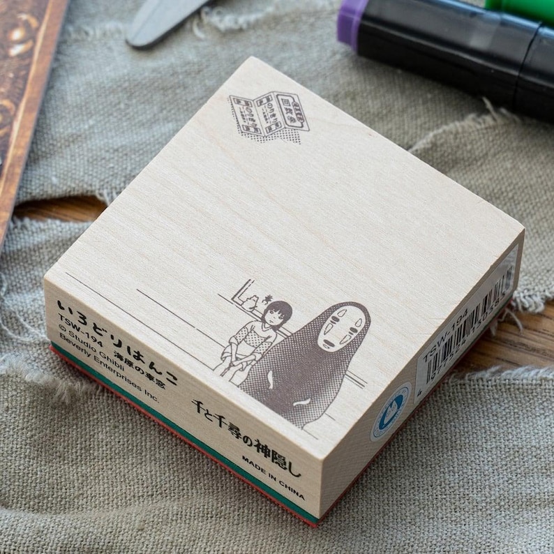 May include: A square wooden stamp featuring a black ink illustration of the characters Chihiro and No-Face from Spirited Away. The stamp includes Japanese text, the code TSW-194, and Studio Ghibli branding printed on the light wood surface.