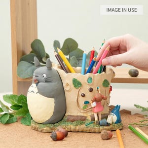 Puede incluir: Un portal&aacute;pices caprichoso con una figura de Totoro gris y un recipiente en forma de tronco de &aacute;rbol. El portal&aacute;pices est&aacute; lleno de l&aacute;pices de colores, y tambi&eacute;n hay una ni&ntilde;a peque&ntilde;a y una figura de Totoro azul. El soporte est&aacute; hecho de un material marr&oacute;n claro.