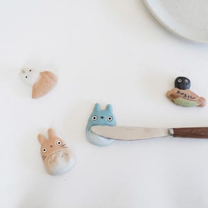 Puede incluir: Cuatro reposa palillos de cer&aacute;mica con forma de personajes de dibujos animados, incluyendo una figura beige, una figura azul que sostiene un cuchillo de acero inoxidable con mango de madera, una figura blanca y una figura oscura con texto japon&eacute;s.