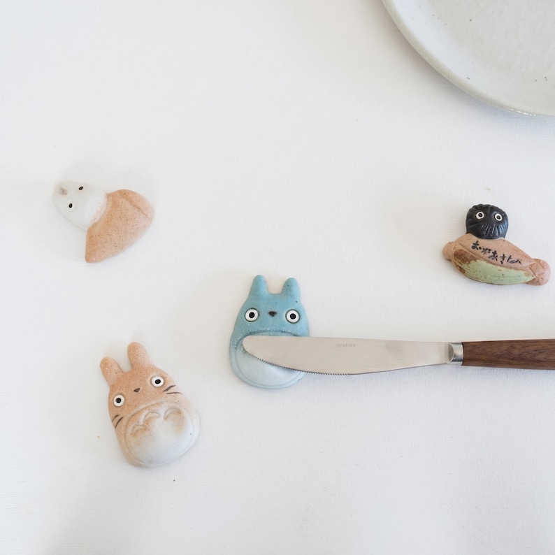 Puede incluir: Cuatro reposa palillos de cer&aacute;mica con forma de personajes de dibujos animados, incluyendo una figura beige, una figura azul que sostiene un cuchillo de acero inoxidable con mango de madera, una figura blanca y una figura oscura con texto japon&eacute;s.