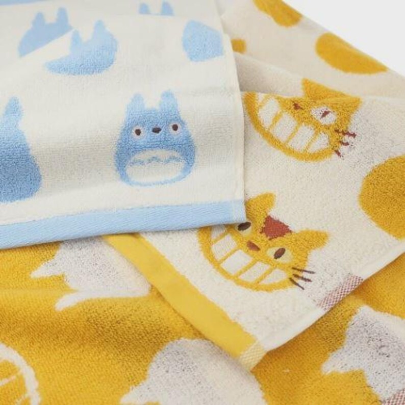 May include: Two folded cotton towels. One is white with blue cartoon characters and a blue border. The other is yellow with a cartoon cat design. The towels are soft and likely for use in a bathroom or kitchen.