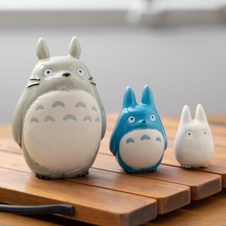 May include: Three ceramic Totoro figurines in varying sizes. The largest is gray with a white belly and dark markings. The middle one is blue, and the smallest is white. All have large eyes and rounded features. They are displayed on a wooden surface.
