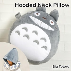 May include: A gray hooded neck pillow shaped like a cartoon character, with a white belly and gray accents. The pillow has a smiling face and is labeled "Big Totoro". A smaller image shows a person wearing a matching hood.