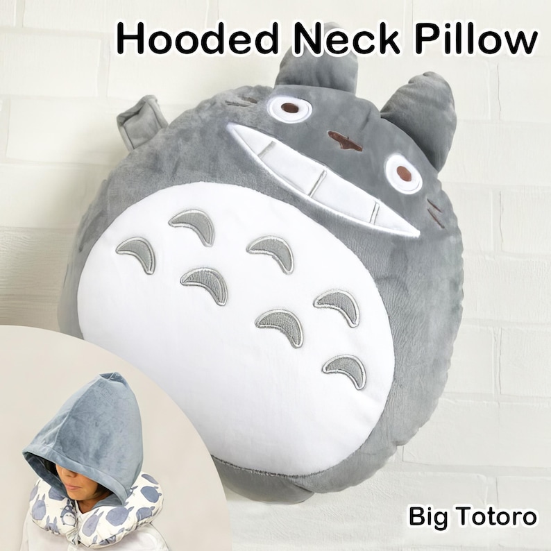 May include: A gray hooded neck pillow shaped like a cartoon character, with a white belly and gray accents. The pillow has a smiling face and is labeled "Big Totoro". A smaller image shows a person wearing a matching hood.
