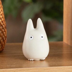 K&ouml;nnte beinhalten: Eine kleine, cremefarbene Keramikfigur, die an Totoro erinnert. Die Figur hat zwei spitze Ohren, schwarze Augen und einen abgerundeten K&ouml;rper. Sie steht auf einem Holzregal, im Hintergrund ist ein geflochtener Korb teilweise zu sehen.