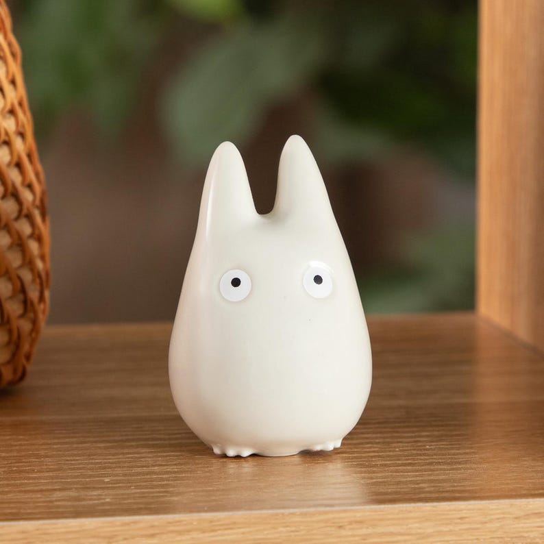 K&ouml;nnte beinhalten: Eine kleine, cremefarbene Keramikfigur, die an Totoro erinnert. Die Figur hat zwei spitze Ohren, schwarze Augen und einen abgerundeten K&ouml;rper. Sie steht auf einem Holzregal, im Hintergrund ist ein geflochtener Korb teilweise zu sehen.