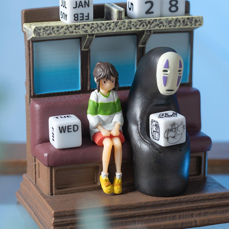 May include: A detailed figurine featuring characters from Spirited Away. The scene includes a seated girl, a figure in a black cloak, and a train car design with a calendar. The figurine has a brown base and a maroon seat.
