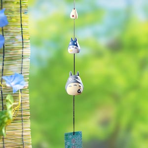 Puede incluir: Un móvil colgante con tres figuras, incluyendo dos que se asemejan a personajes de Totoro, en gris, blanco y azul. El móvil tiene una pequeña etiqueta con caracteres japoneses. El fondo es verde borroso.