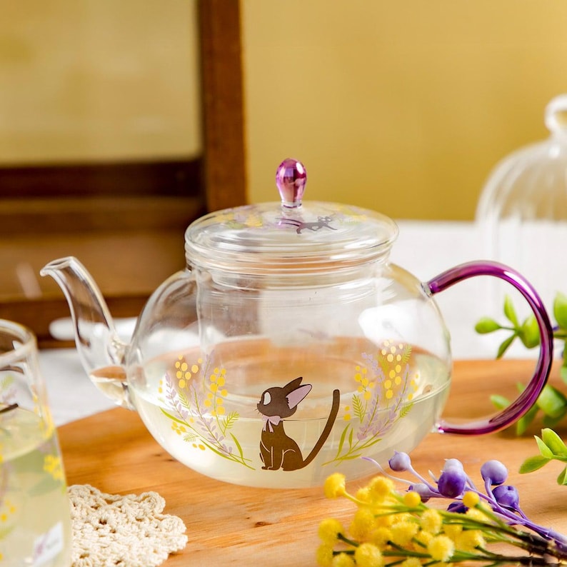 Puede incluir: Una tetera de cristal transparente con un gr&aacute;fico de un gato negro con un lazo rosa, rodeado de ilustraciones florales amarillas y moradas. La tetera tiene un asa morada y un pomo morado a juego en la tapa, con un pico curvo para servir.