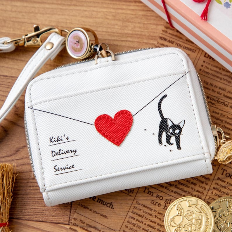 May include: White wallet with a zipper closure, featuring a red heart and a black cat illustration. The wallet has a wrist strap and the text "Kiki's Delivery Service". A gold-colored charm is attached to the zipper.