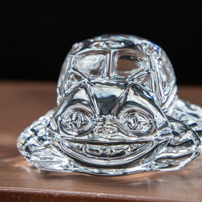 May include: A clear, glass turtle figurine with a smiling face. The turtle's shell is detailed with geometric patterns, and its eyes and mouth are clearly defined. The figurine is sitting on a brown surface.