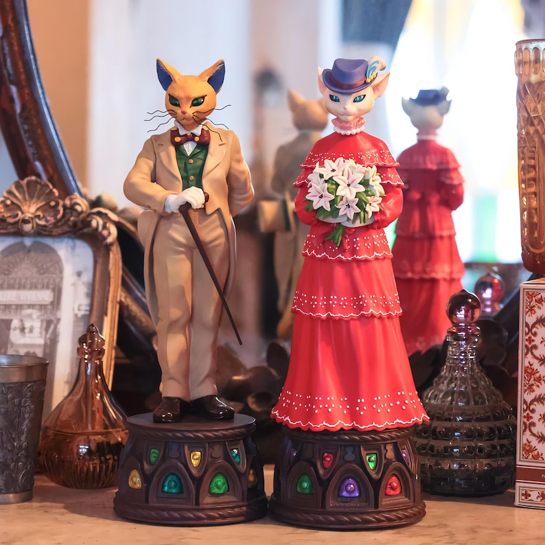 Puede incluir: Dos figuras de gato, una con traje beige y bast&oacute;n, la otra con vestido rojo y flores. Ambas sobre pedestales decorativos con detalles coloridos tipo vidriera. Un espejo refleja las figuras.