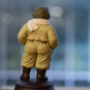 May include: A ceramic figurine of a pilot, viewed from the back. The pilot wears a yellow jumpsuit, brown boots, and a green helmet with goggles. A white scarf is draped around the neck. The figurine is standing on a brown base.