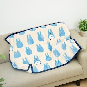 May include: A cream-colored blanket with a pattern of blue cartoon characters. The blanket has a dark blue border and is draped over a beige sofa. The blanket is soft and plush, perfect for snuggling up with.