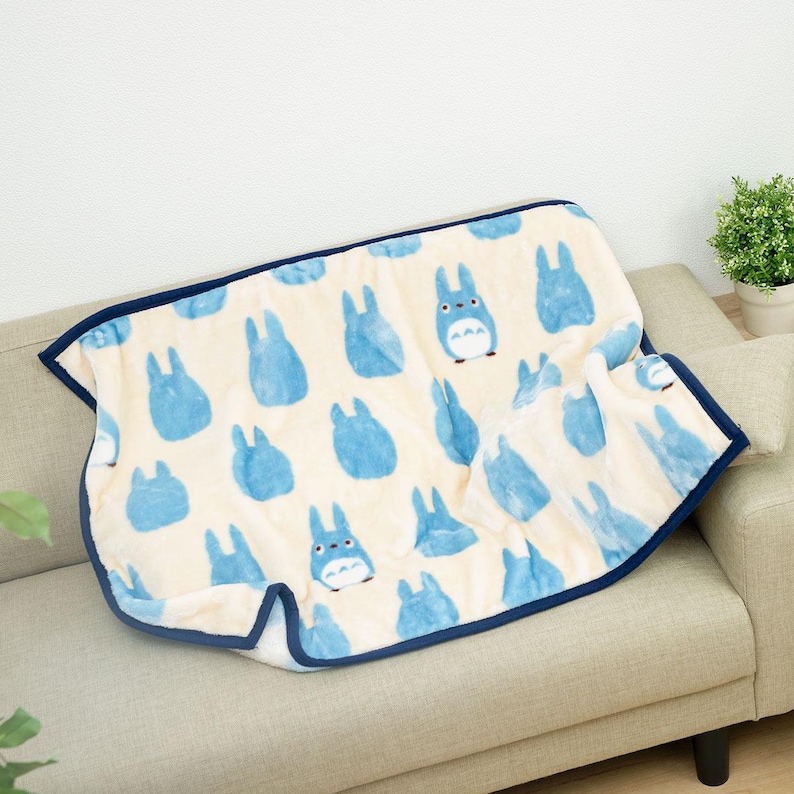 May include: A cream-colored blanket with a pattern of blue cartoon characters. The blanket has a dark blue border and is draped over a beige sofa. The blanket is soft and plush, perfect for snuggling up with.