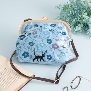 May include: A light blue crossbody bag with a floral and cat embroidery design. The bag has a brown strap and a gold-coloured clasp. The bag is decorated with blue and purple flowers and black cat silhouettes.