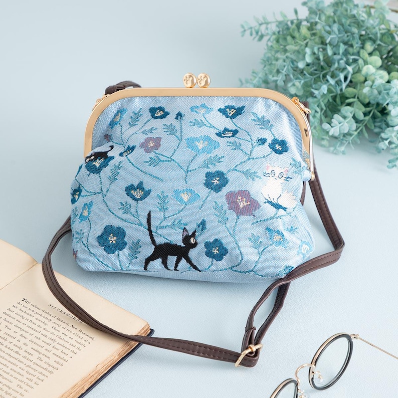 May include: A light blue crossbody bag with a floral and cat embroidery design. The bag features a brown strap and a gold-colored clasp. The bag is decorated with blue and purple flowers and black cat silhouettes.