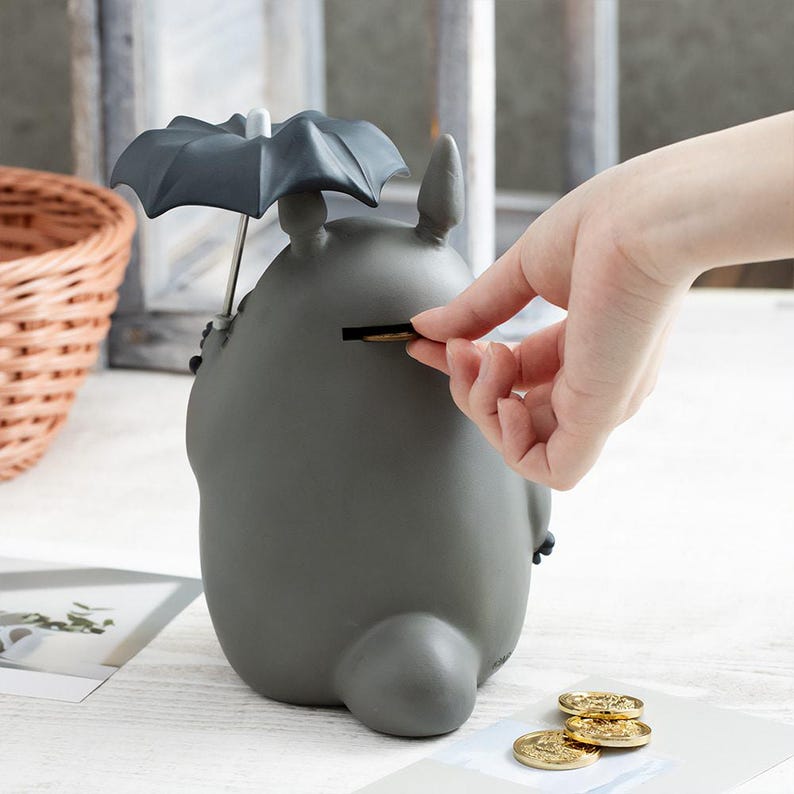 Puede incluir: Una hucha de cer&aacute;mica gris mate con forma de criatura redonda y orejas puntiagudas, que sostiene un paraguas gris oscuro. Una ranura para monedas se encuentra en la parte posterior de la figura. Tres monedas doradas est&aacute;n cerca de la base.