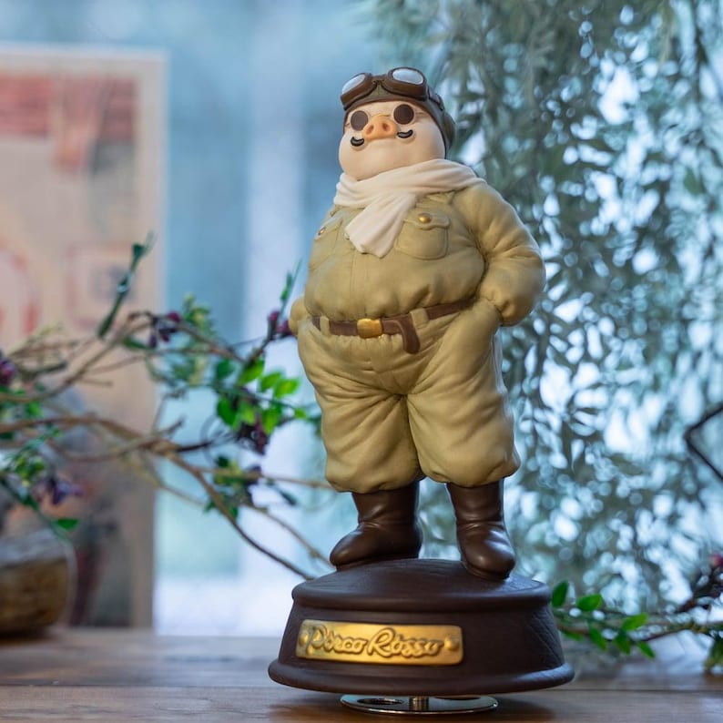 May include: A figurine of a pig in a pilot's outfit, complete with goggles, a scarf, and a khaki jumpsuit. The figure stands on a brown base with the gold text "Porco Rosso". The theme is aviation and the figurine is likely a collectible.