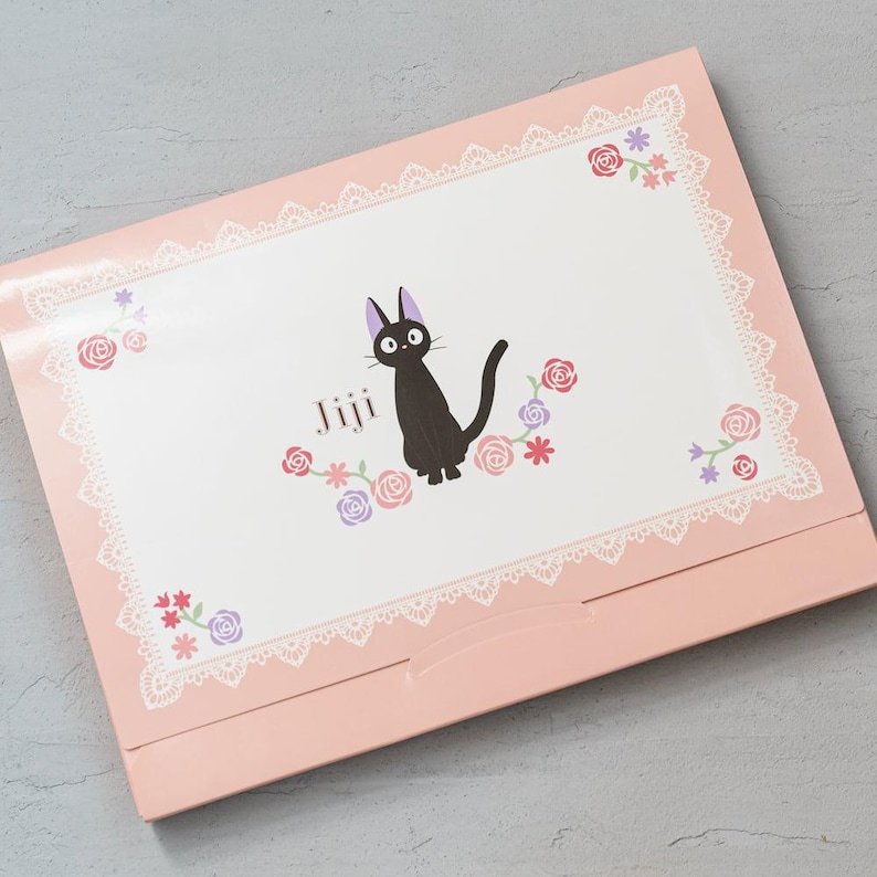 May include: A pink box with a black cat illustration and the name "Jiji". The design includes floral accents and a lace border. The box is closed and has a light pink colour. The overall design is cute and decorative.
