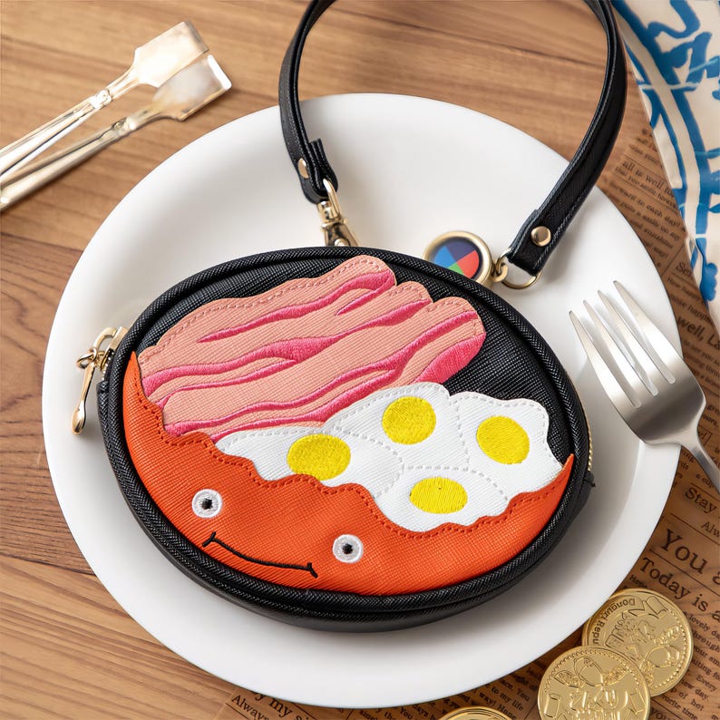 May include: A circular black purse with a breakfast design, featuring embroidered bacon, fried eggs, and a smiling orange face. It has a black handle and gold zip. The purse is on a white plate.