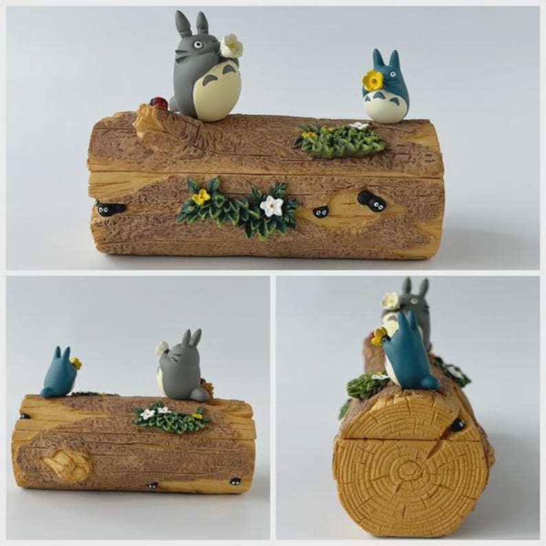 May include: A decorative wooden box featuring Totoro characters. The box is shaped like a log with carved details, adorned with small flowers, greenery, and Totoro figures in gray and blue. The box has a light brown color.