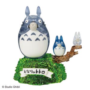 Op de afbeelding: Een doorschijnende blauw-witte Totoro-figuur met twee kleinere figuren op een bruine tak, geplaatst op een groene basis. De basis heeft een witte banner met Japanse tekst. De figuren zijn van de Studio Ghibli-collectie.